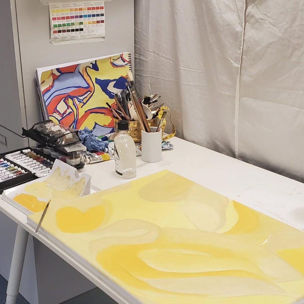 Art desk with a yellow abstract painting, brushes, paints, and a colorful artwork in the background.